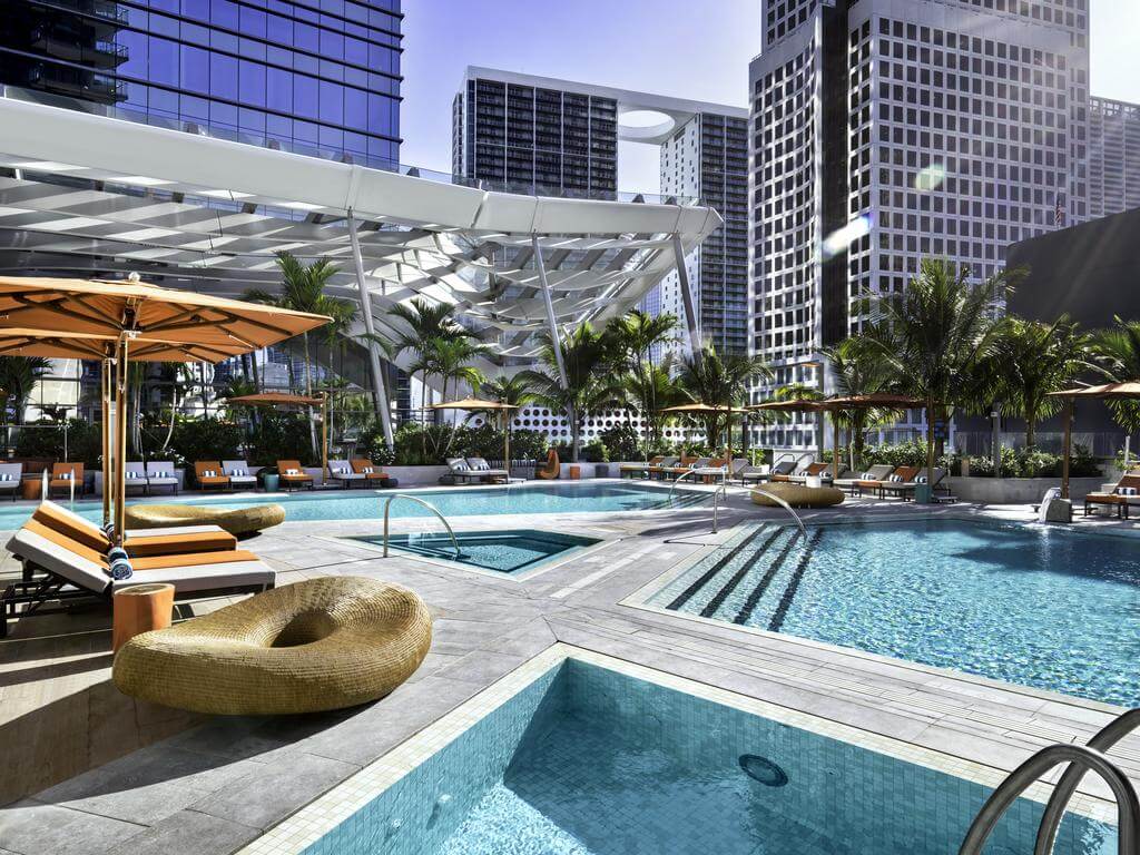 A luxurious rooftop pool deck with multiple pools, lounge chairs, and umbrellas, surrounded by modern skyscrapers and palm trees, creating a vibrant urban oasis.