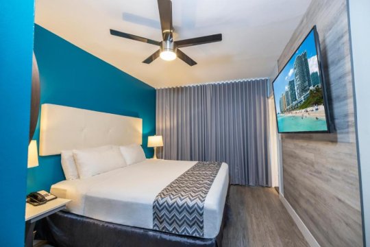 Modern hotel room with a king-size bed, bright blue accent wall, large flat-screen TV displaying a beach scene, and gray curtains.