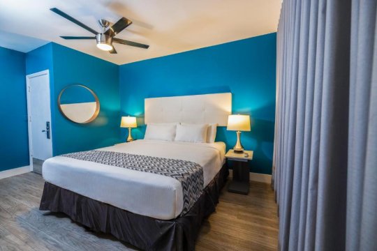Modern hotel room with a king-size bed featuring a white headboard, blue walls, and a ceiling fan. A round mirror and bedside lamps add to the contemporary hotel decor.