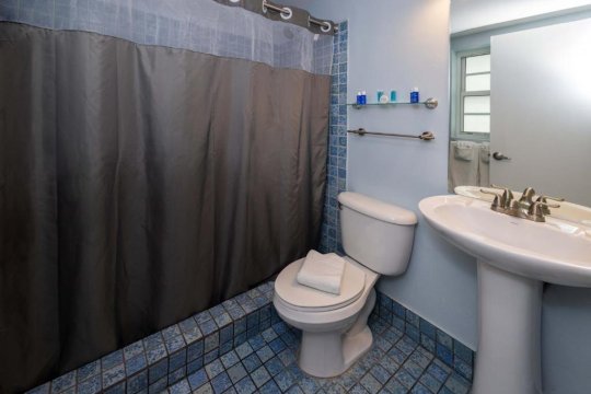 A small, clean bathroom with blue tiled floor and walls, featuring a white toilet with a folded towel, a white pedestal sink with chrome faucet, and a grey shower curtain.