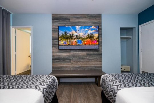 Modern hotel room with two beds, a flat-screen TV displaying a tropical beach scene, and wood-paneled accent wall.
