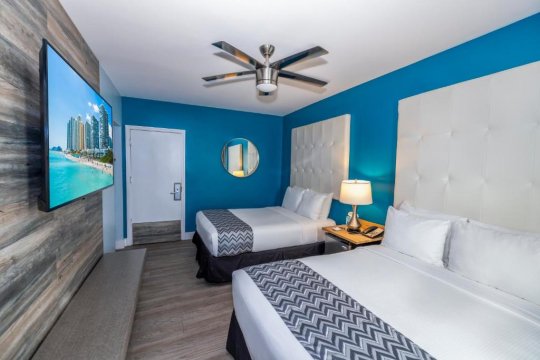 A hotel room with two queen beds, blue walls, and a large flat-screen TV mounted on a wood-paneled wall.