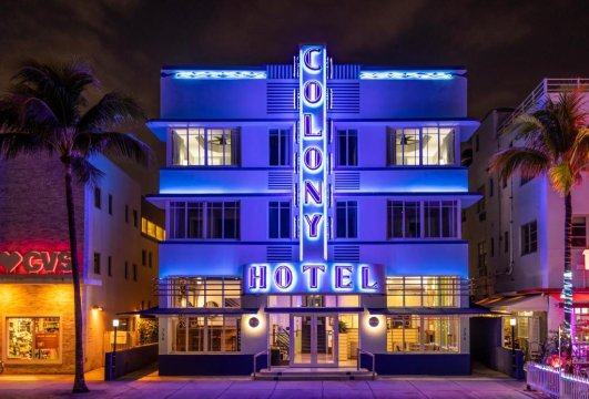 The Colony Hotel in Miami Beach glows with vibrant blue neon lights at night, its Art Deco facade illuminated against the dark sky. Palm trees frame the scene, hinting at a tropical getaway.