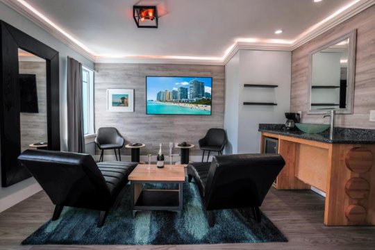 Luxurious hotel suite living area with two black leather recliners facing a coffee table, a large flat-screen TV displaying a beach scene, and a wet bar with a mini-fridge and coffee maker.