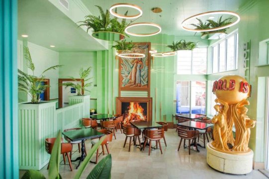 A stylish, mint-green dining room with a fireplace, gold statue spelling "WORLD IS", and hanging plants. Modern, art deco interior design.