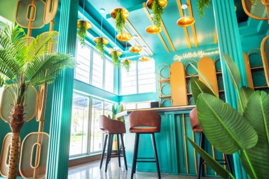 Vibrant turquoise interior of a modern cafe with a bar, featuring hanging plants, gold pendant lights, and brown leather bar stools. "Say Hello to My..." neon sign visible.