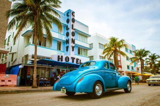 A vintage blue car drives past the iconic Colony Hotel in Miami Beach, with palm trees lining the street and other classic cars visible.