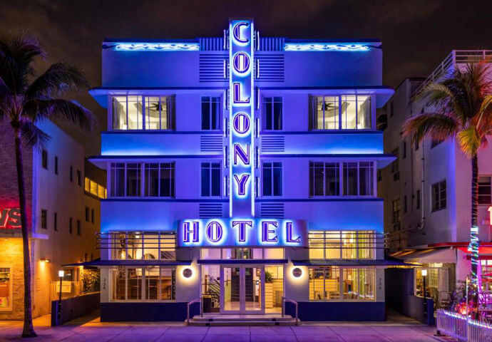 Colony Hotel, Ocean Drive, Miami Beach at night — iconic blue neon Art Deco facade with vertical "COLONY / HOTEL" signage, palm trees flanking the entrance.