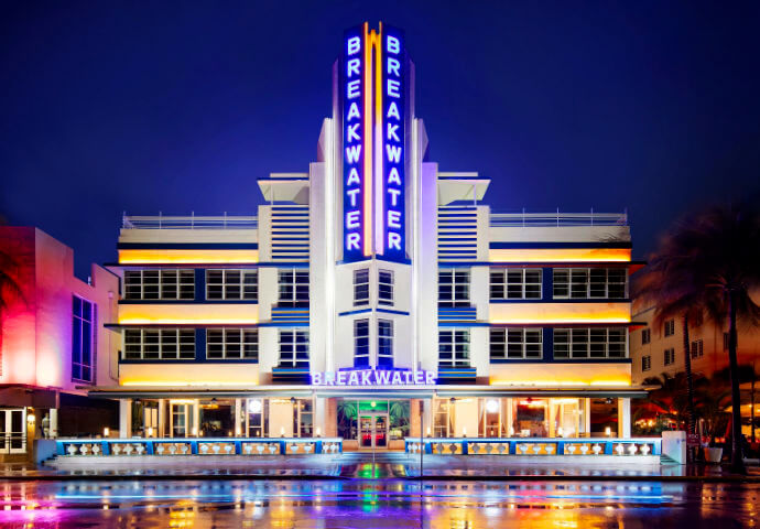 The Breakwater Hotel, Ocean Drive, Miami Beach at night — symmetrical Art Deco facade with vertical blue neon "BREAKWATER" tower sign, wet street reflecting the lights.