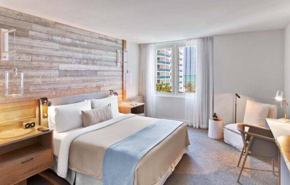 A modern hotel bedroom with a king-size bed, light wood accent wall, and a large window overlooking a beach and ocean view. The room features a comfortable seating area with a desk and chair.