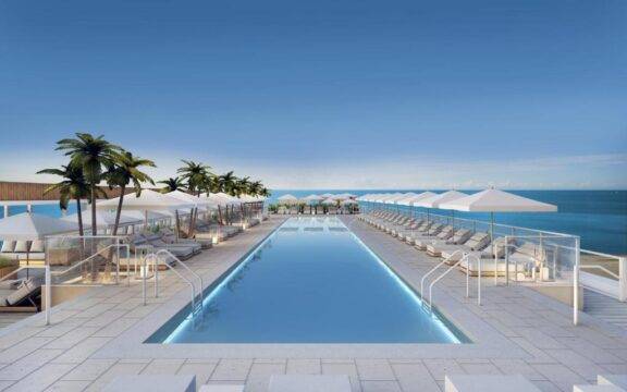A luxurious resort infinity pool with bright blue water and underwater lighting, surrounded by white lounge chairs and umbrellas, overlooks a serene ocean horizon with palm trees lining the deck.