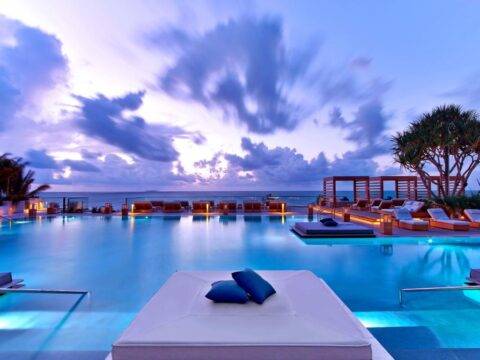Luxury hotel pool at dusk with a vibrant purple and orange sky over the ocean, featuring comfortable lounge beds and ambient lighting.