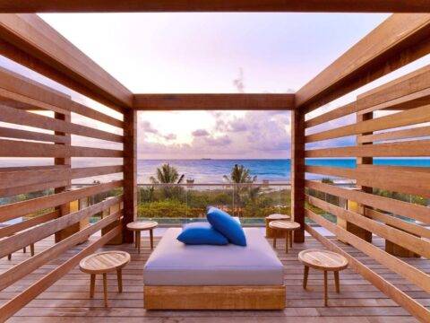 A tranquil wooden cabana with a large daybed and blue pillows offers a stunning ocean view at sunset, featuring palm trees and a serene coastal landscape.