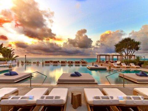 Luxury resort pool at sunset with plush lounge beds and umbrellas, overlooking a calm ocean with dramatic, colorful clouds.