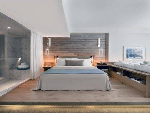 Luxurious hotel bedroom with a king-size bed, modern wood-paneled wall, and a sleek glass-enclosed shower. Natural light illuminates the serene, upscale interior.