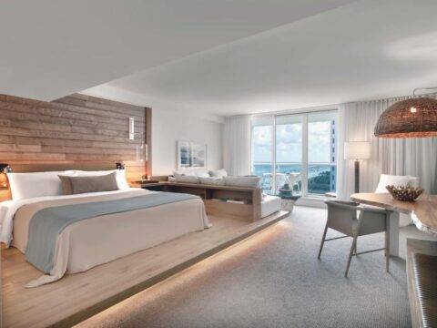 Luxurious hotel suite with a king-size bed, plush seating area, and balcony overlooking the ocean, featuring natural wood accents and modern decor.