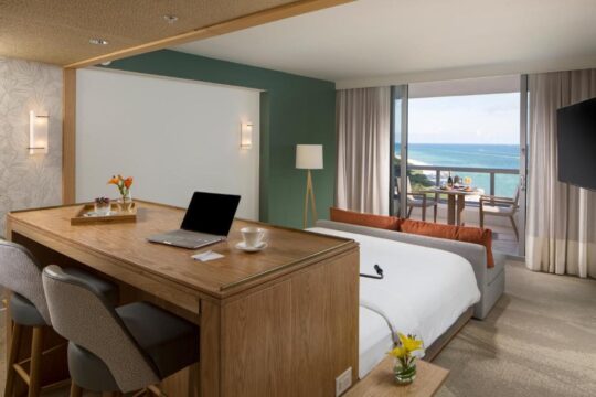 Modern hotel room with a desk and bed facing a balcony with ocean views, featuring a laptop, coffee cup, and orange accents.