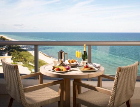 A romantic breakfast for two on a balcony overlooking the ocean, featuring champagne, fresh fruit, pastries, and coffee, with two chairs set at a table.