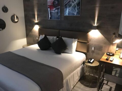 Modern hotel room with a king-size bed, plush pillows, and bedside tables featuring lamps and a phone. The wall behind the bed has a textured wood paneling and features framed art.