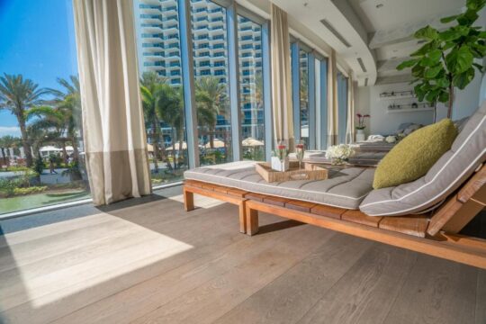 Luxury spa relaxation lounge with chaise lounges, plush cushions, and a tray with champagne glasses and flowers, overlooking a tropical resort pool and modern high-rise buildings.