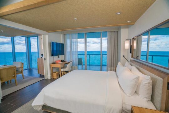 Luxurious hotel room with a king-size bed, desk, and floor-to-ceiling windows offering a breathtaking ocean view. This serene oceanfront hotel room features modern decor and expansive sea vistas.