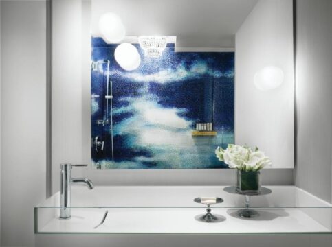 Modern bathroom vanity with a clear glass countertop, sleek chrome faucet, and a vibrant blue mosaic tile shower wall featuring a cloudy sky design. A small vase of white flowers adds a touch of elegance.