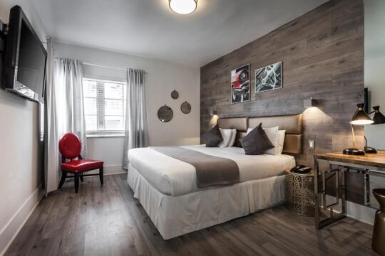 Modern hotel bedroom with a king-size bed, grey wood accent wall, red chair, and desk.