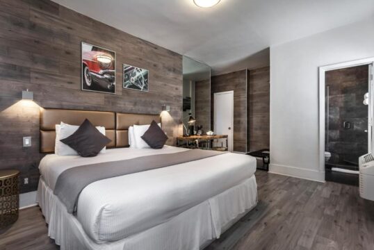 Modern hotel room with a king-size bed, wood-paneled accent wall, and a sleek en-suite bathroom. Artwork of a classic red car adds a touch of retro style.
