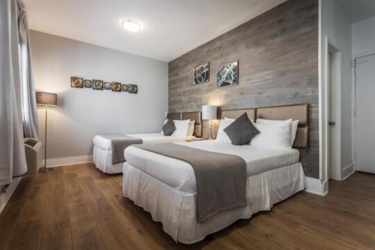 A modern hotel room with two queen beds, featuring a rustic wood-paneled accent wall, grey bedding, and a contemporary floor lamp.