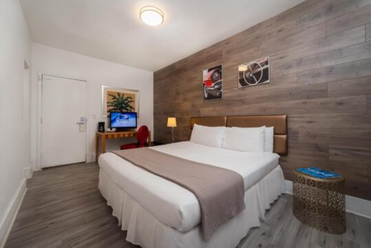 Modern hotel room with a king-size bed, wooden accent wall, and vintage car artwork. A desk with a TV and a red chair are visible.
