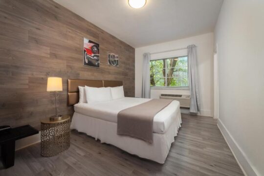 A cozy hotel room features a king-size bed with white linens and a grey throw, a wooden accent wall with vintage car art, and a bedside lamp on an ornate table.