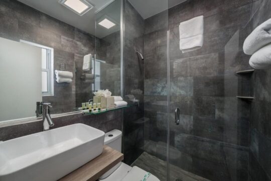 Modern hotel bathroom with a sleek white vessel sink, dark tiled walls, and a glass-enclosed shower with fluffy white towels.