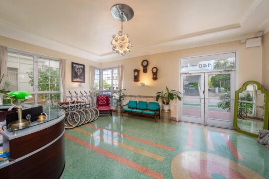 Art Deco lobby with vintage bicycles, a red armchair, teal bench, and a terrazzo floor with colorful geometric patterns.