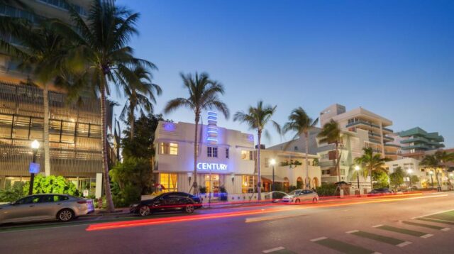 Art Deco hotel "Century" in Miami Beach at dusk, with palm trees, cars, and light trails from traffic along Ocean Drive.