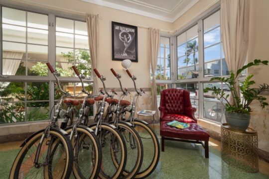 A line of vintage bicycles with chrome handlebars and brown seats sits in a bright, airy room with large windows, a red tufted chaise lounge, and lush green plants. An "Art Deco Weekend" poster hangs on the wall.