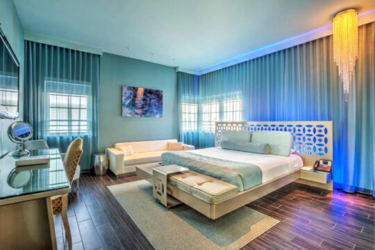 Luxurious hotel suite with a king-size bed, blue accent lighting, and a modern chandelier. A white sofa and desk with a makeup mirror are also visible.
