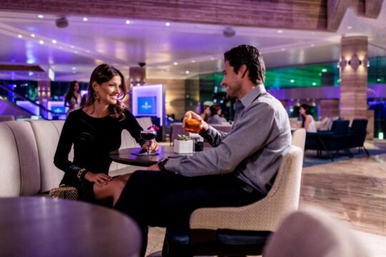 A couple enjoys drinks at a dimly lit, upscale lounge bar, with purple ambient lighting creating a sophisticated atmosphere. The woman, in a black dress, smiles as she holds a wine glass, while the man in a grey shirt sips from his cocktail, seated at a plush armchair.