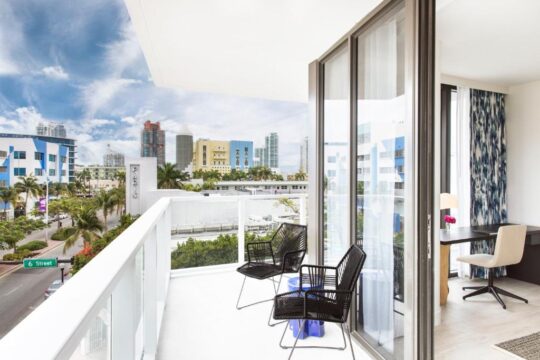 Balcony overlooking Miami Beach with colorful buildings and palm trees, featuring modern chairs and a city view.