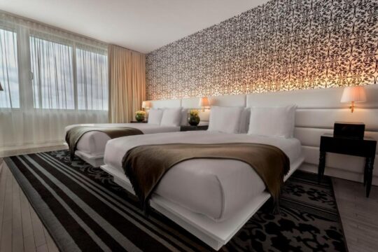 A modern hotel room features two queen beds with crisp white linens and plush headboards, accented by a rich brown throw blanket draped across each. A striking black and white patterned wallpaper adorns the wall behind the beds, complemented by elegant bedside lamps.
