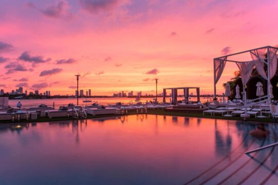 Luxury poolside lounge at sunset with a vibrant pink and orange sky reflected in the calm water, overlooking a city skyline.