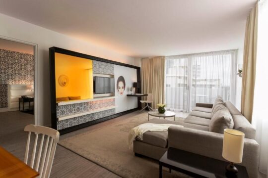 A modern hotel suite living room with a large sectional sofa, a sleek entertainment unit featuring a TV and decorative panels, and large windows with sheer curtains.