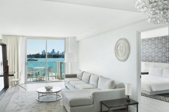 Spacious white living room with a large sectional sofa and a balcony overlooking a sparkling blue bay and city skyline. The room features a modern chandelier and a glimpse of a bedroom with a patterned wall.