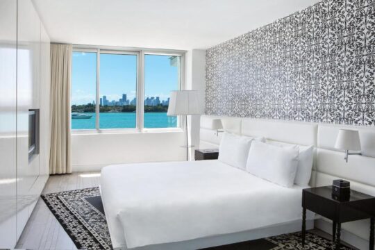 Modern white hotel bedroom with a king-size bed, black and white patterned wallpaper, and a large window overlooking a bright blue bay with city skyline and a yacht.