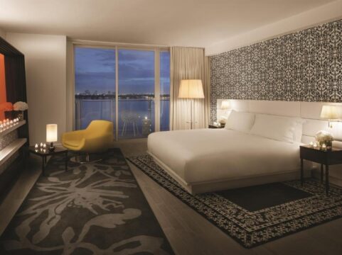 Luxury hotel bedroom with a king-size bed, modern armchair, and a large window overlooking a city skyline at dusk.