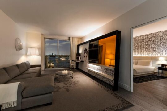 Luxurious hotel suite living room with a large grey sectional sofa, a glass coffee table, and a striking mirrored wall unit featuring a TV and decorative shelving, opening to a bedroom with a king-size bed.