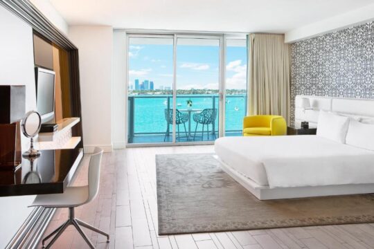 Modern hotel room with a king-size bed, desk, and balcony overlooking a bright blue ocean with city skyline in the distance.
