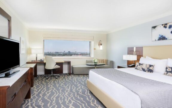 A spacious hotel room with a king-size bed, a work desk, and a large window offering a city view. The room features a grey patterned carpet, a modern sofa, and a television.