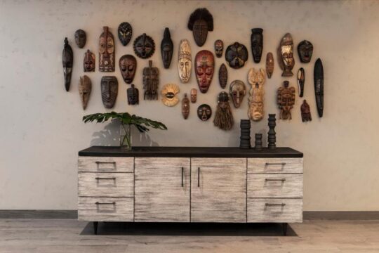 A wall adorned with a diverse collection of African tribal masks, ranging in size and intricate detail, above a rustic wooden sideboard with drawers.