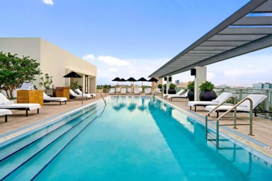 Rooftop pool with lounge chairs and umbrellas, offering a clear blue sky and city views. This resort amenity is perfect for relaxation and enjoying the sun.