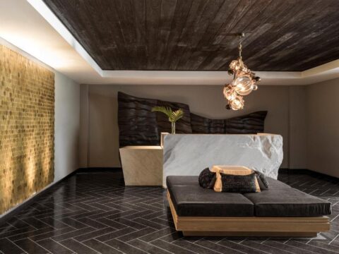 Luxurious spa reception area with a large white stone reception desk, dark wood ceiling, and a textured gold wall panel. A dark grey daybed with a patterned cushion sits in the foreground.